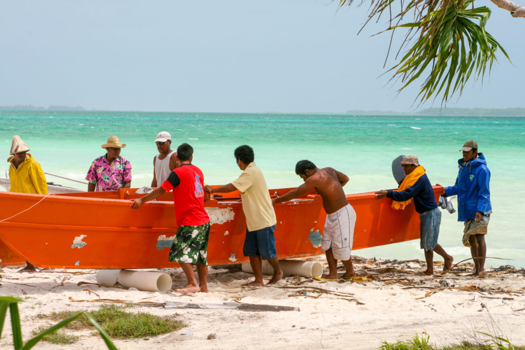 Eight men are rolling an orange boat onto the shore from the ocean.